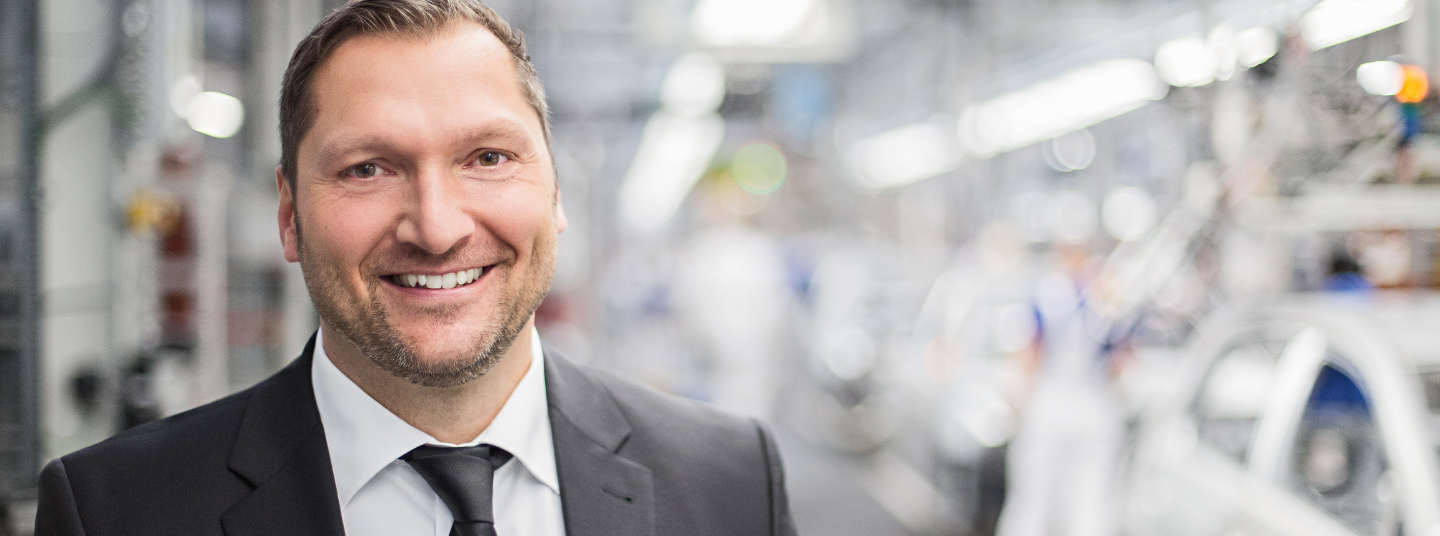 Ein Ingenieur im Einsatz bei unserem Kunden in der Automobilindustrie. Er lächelt in die Kamera und steht in einer Fertigungshalle.